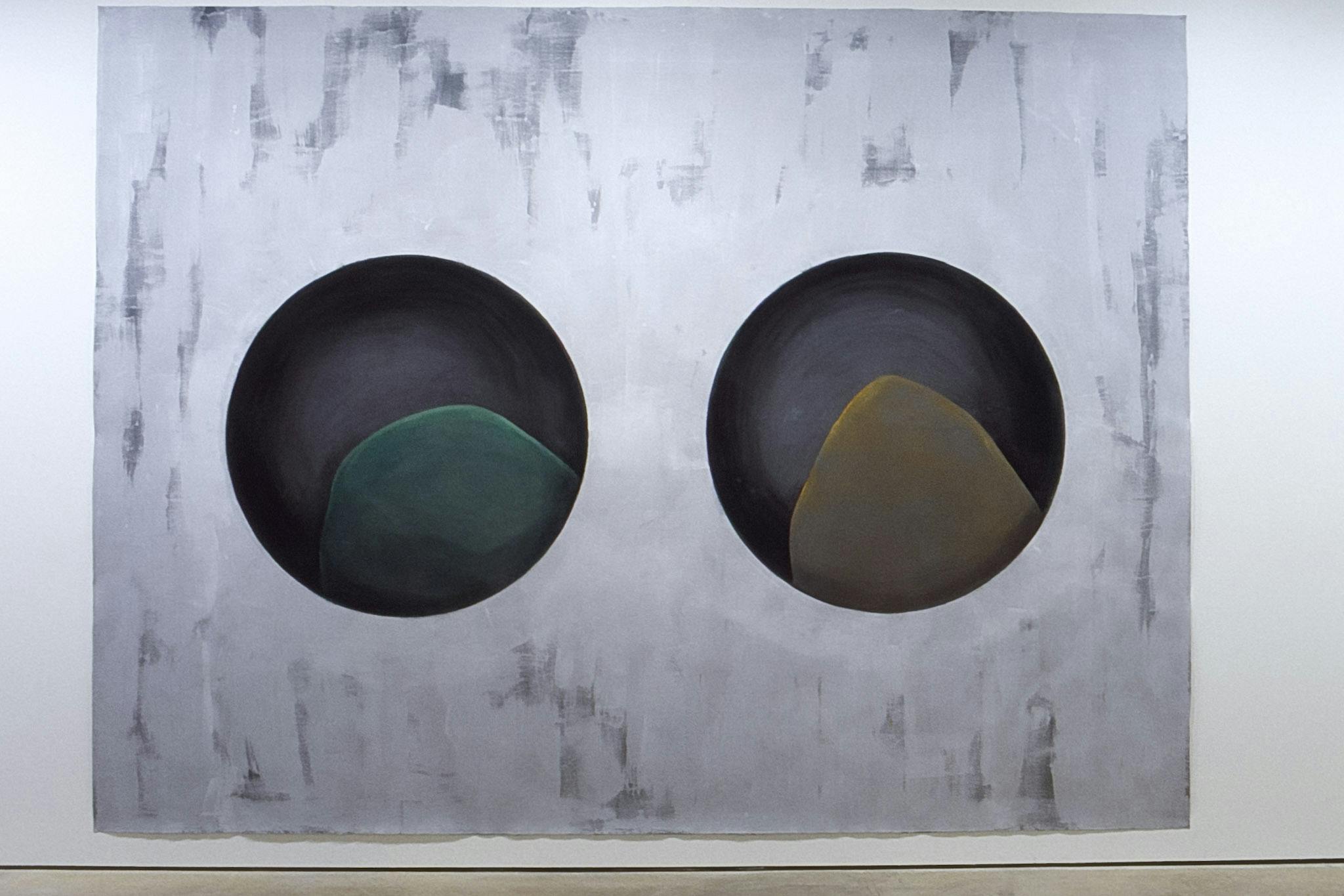 A large painting is installed on a gallery wall. Two black circles in the middle, which have organic shapes inside, are depicted in the light-gray background resembling a surface of a marble slab. 