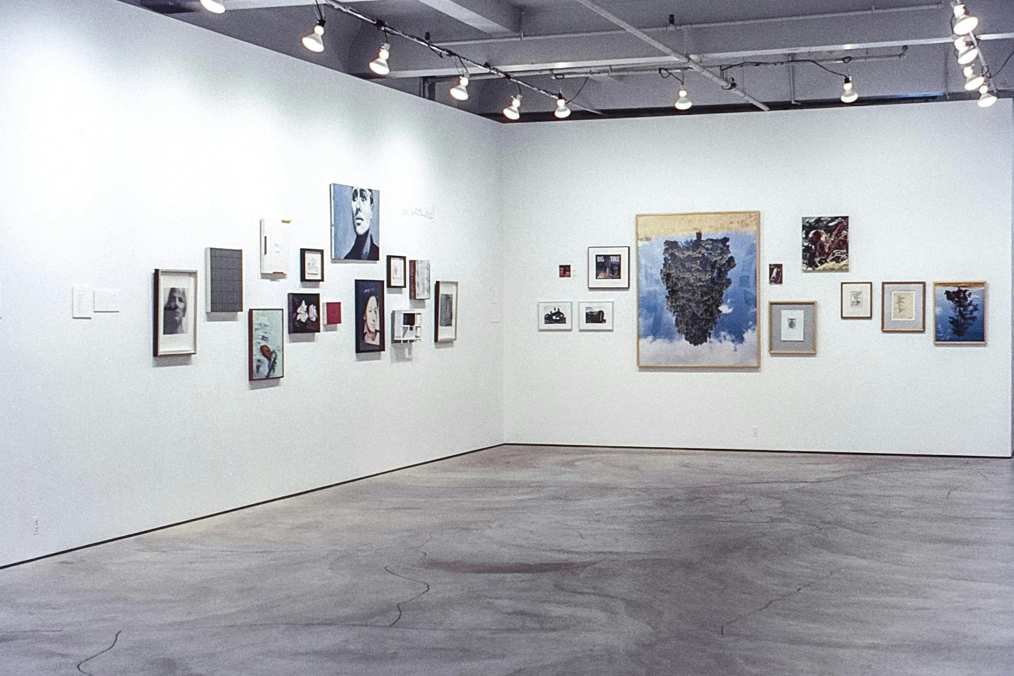 A corner of a gallery space with several artworks mounted across both walls. The works vary in size, framed and unframed. On the right, there is a large photo of a tree with a blue sky, upside down.