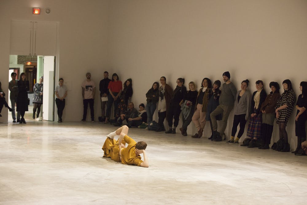 A performer dressed in yellow lays on the floor of a gallery space with audience members standing around the perimeter. They are laying on their side, propping their head up with their arm. 