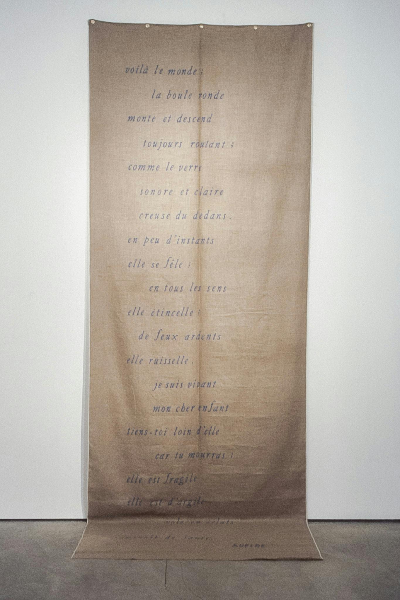 Mounted on a white wall, a long piece of brown fabric drapes to the floor. The fabric has 22 french phrases on it in italicized, purple lowercase text. The first phrase reads: "voilà le monde."