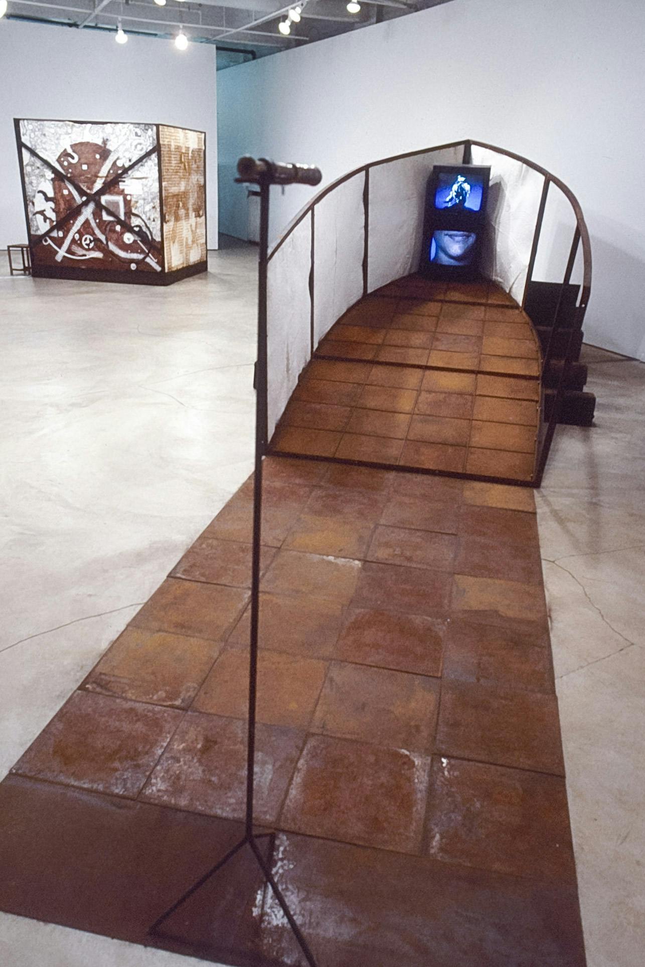 Two large sculptures are installed in the gallery. A white boat head with wood tile flooring is placed in the front. Two CRT TVs are placed on the boat. A cubical-shaped sculpture is in the back.