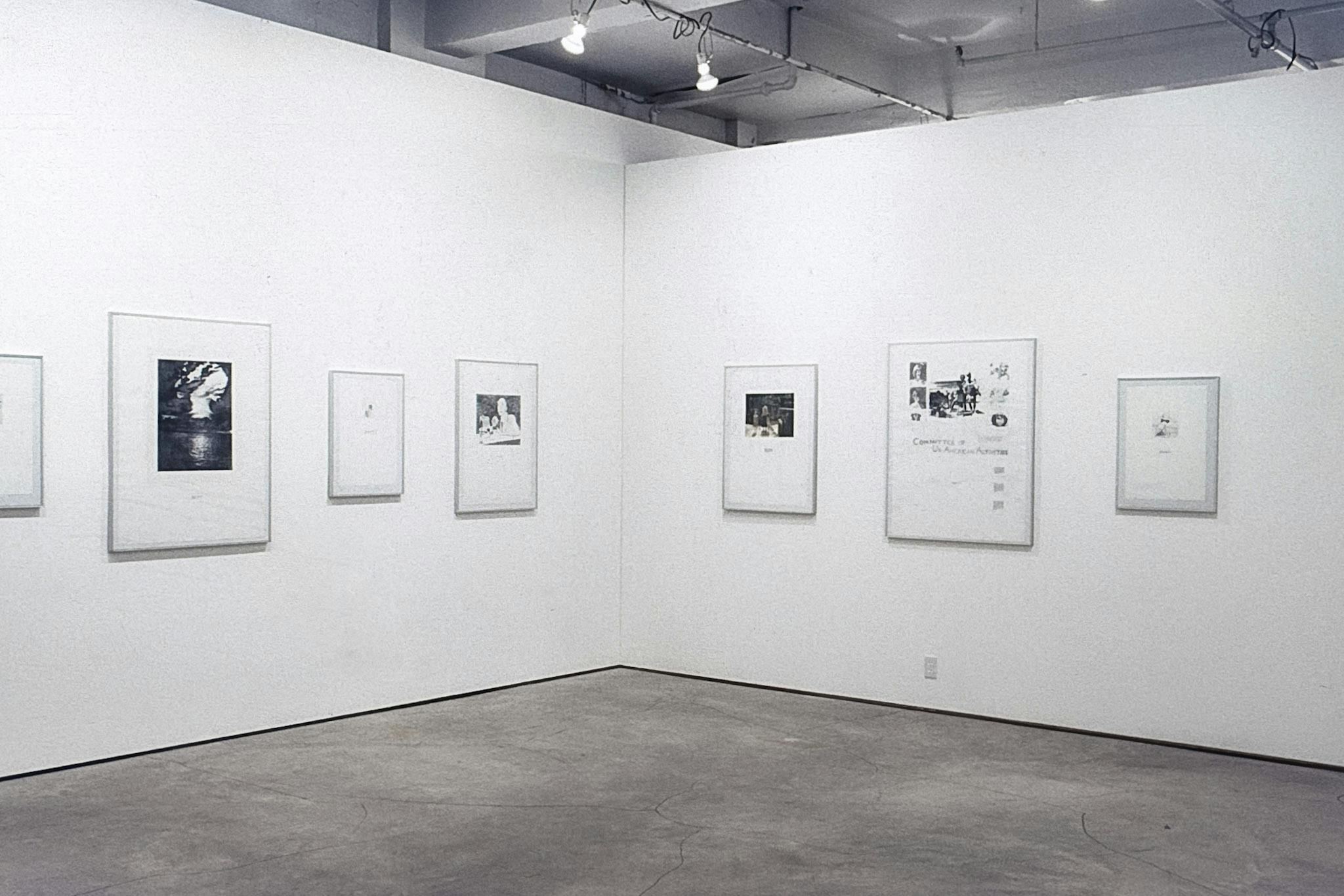 The corner of a gallery space. Different-sized illustrations and collages are in metal frames mounted on the white walls.