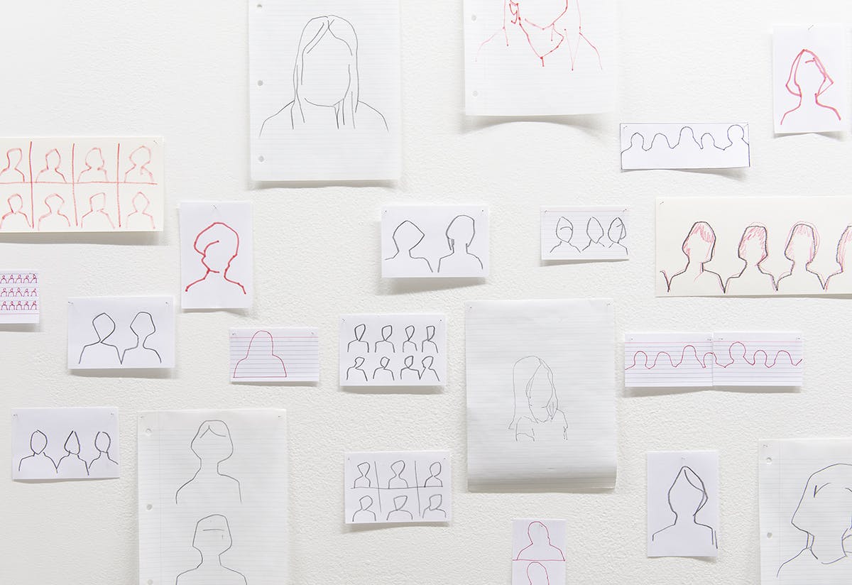 This is a close-up view of Faye HeavyShield’s drawings installed on a gallery wall. They were drawn with several different mediums, including markers and pens. They depict the shapes of human heads.