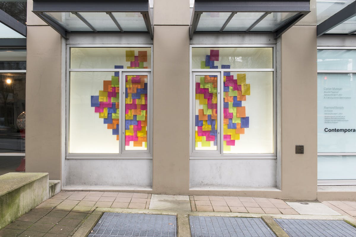 Dozens of coloured sticky notes mounted in a window on CAG’s exterior facade. Random combinations of semi-transparent Roman letters are visible on these overlapping pieces of paper.