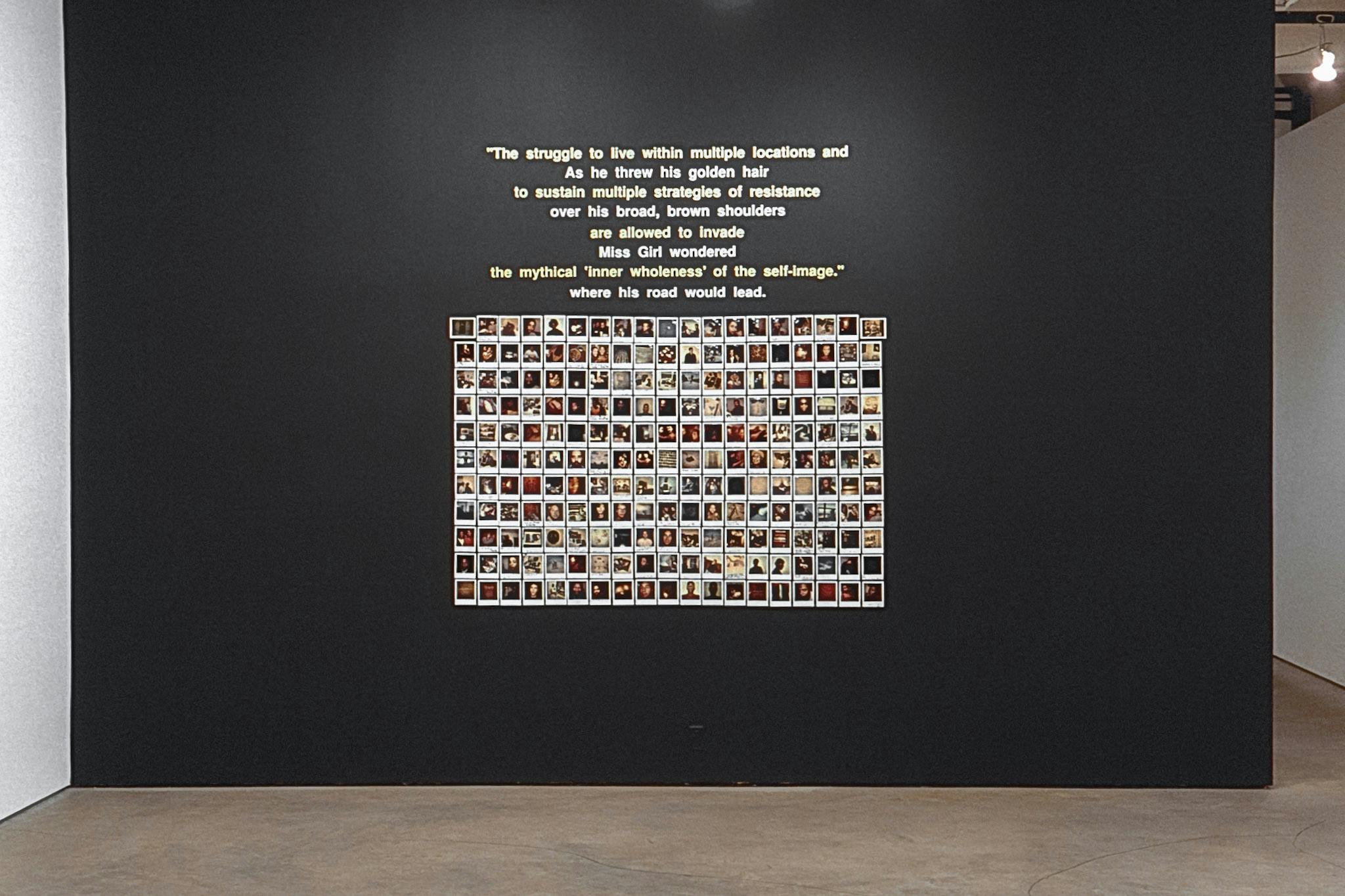 A black wall in a gallery with text and dozens of Polaroid photos. The text is composed of two different excerpts alternating, one in yellow and one in white, intertwining in a disorienting way.