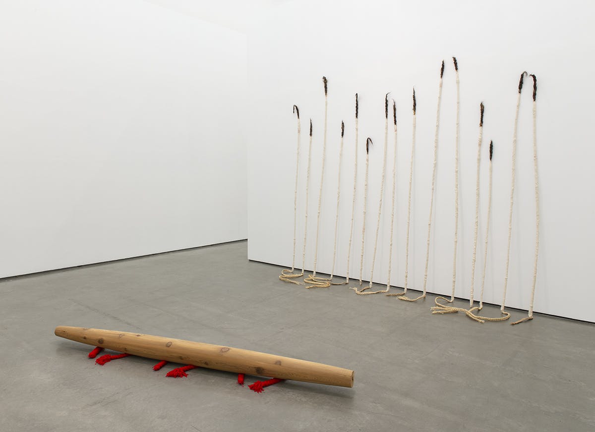 This is an installation shot of Charlene Vickers’ solo exhibition. A large bead-shaped sculpture made of wood sits on the floor. Behind it, a series of spear-shaped sculptures lean against the wall. 
