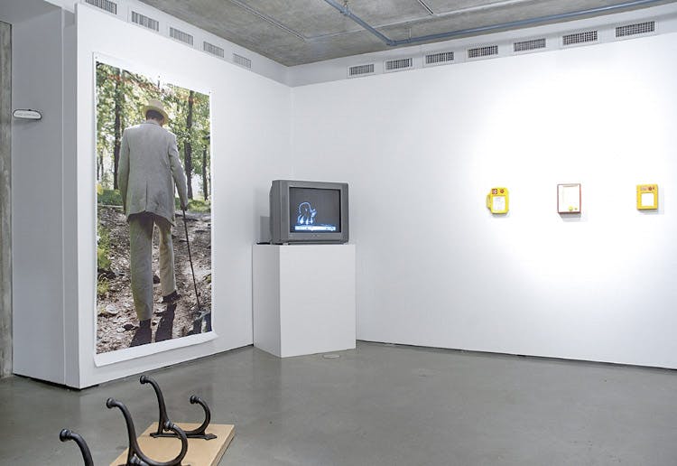 An installation image of Daniel Olson’s art exhibition at CAG. A large poster of a person in a grey suit is mounted on a wall. A CRT TV is placed on a pedestal at the corner of a gallery.