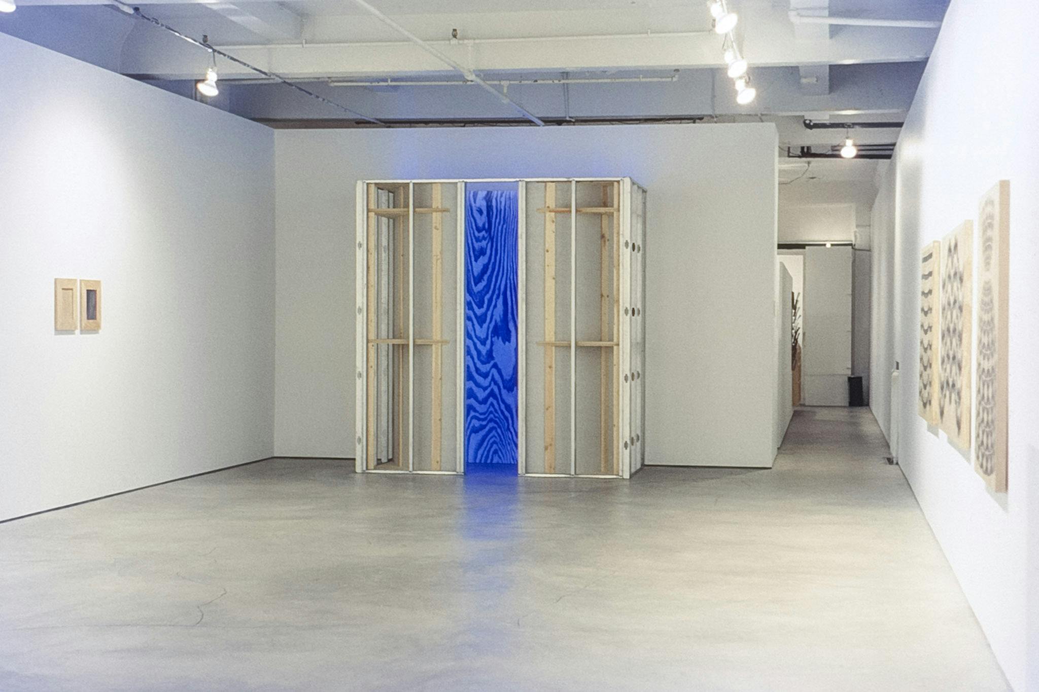A gallery space with artworks on the walls. The work in the centre is a large sculpture made of a wooden frame. The opening of the structure shows a large wood-pattern lit in bright blue.