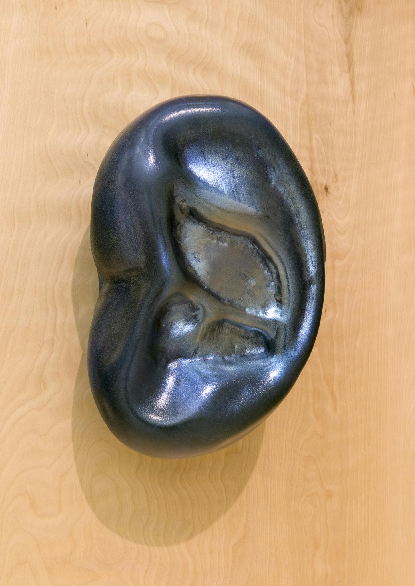Image of a dark, metallic coloured sculpture on a wooden surface. The sculpture resembles the form of a human ear. 
