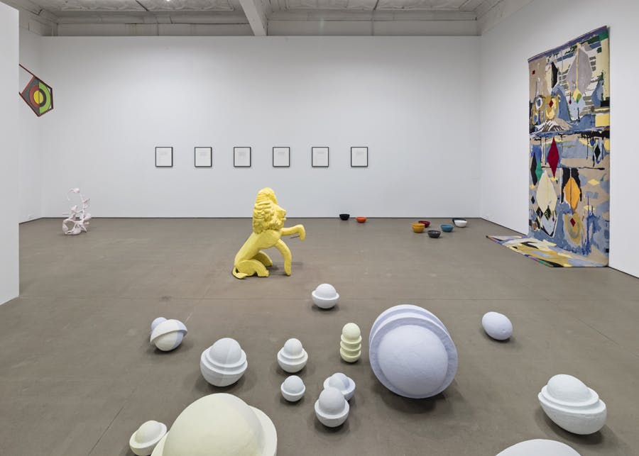 Many artworks are installed in a gallery space. A yellow lion sculpture and various sized ceramic sculptures are placed on the floor. A rug and other two-dimensional pieces are installed on the walls. 