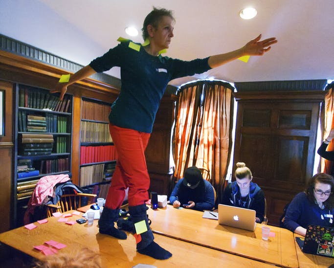 Image of Margaret Dragu performing on a big table in front of book-filled bookshelves. Dragu looks straight ahead, reaching one arm to the front and another to the back. Yellow sticky notes are attached to Dragu’s arms. 