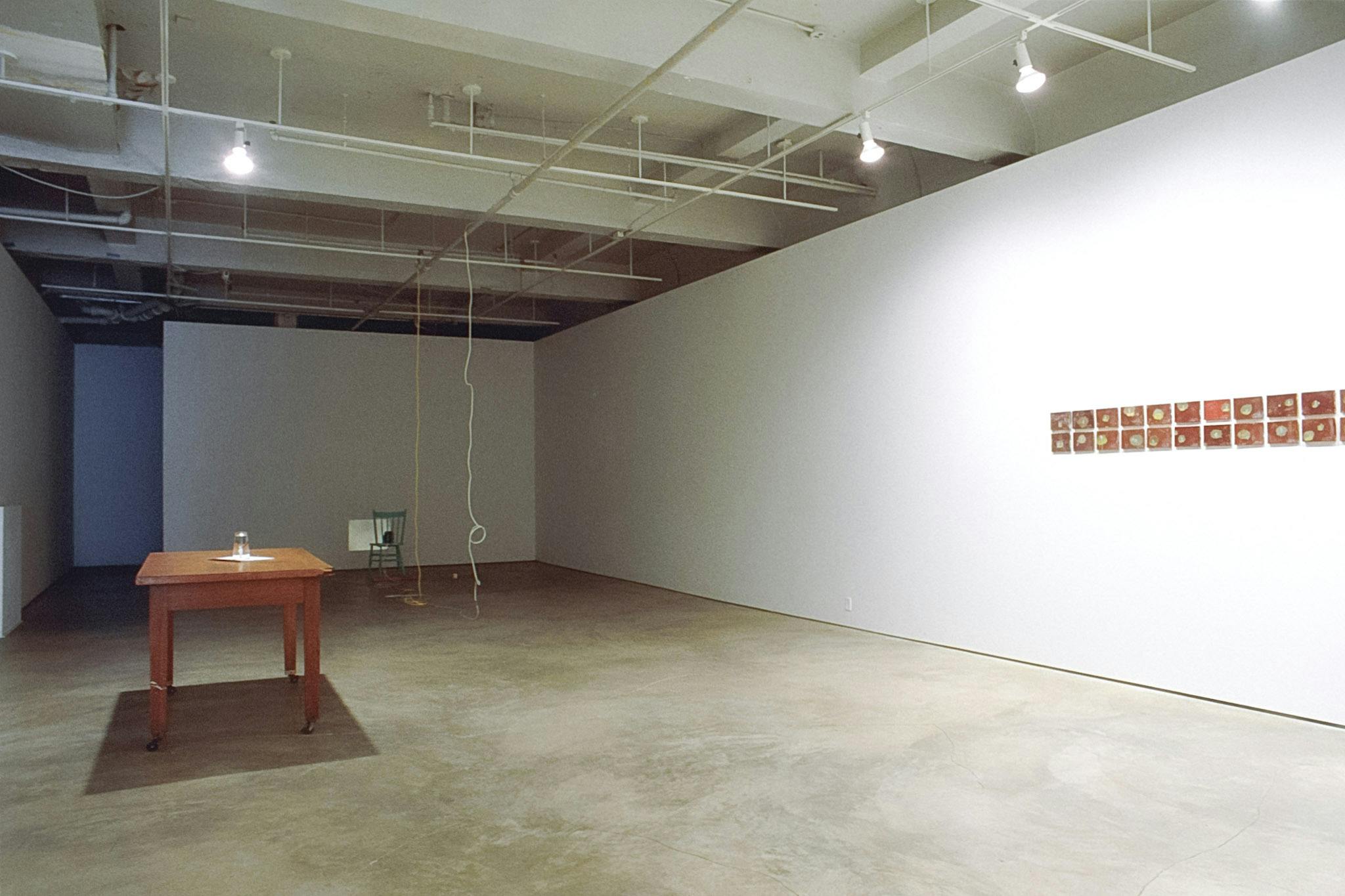 A wooden desk and small wall tiles are installed in the front space of the gallery. in the back, white and yellow strings are hanging from the ceiling in front of a painted wooden chair on the floor. 