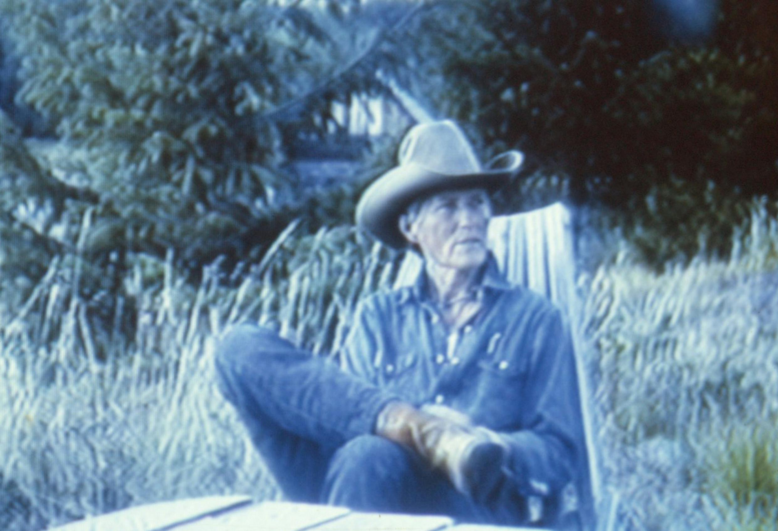 A blue-toned video still of a person sitting in a chair outside with tall grass and trees around them. They are wearing a denim shirt and a cowboy hat. 