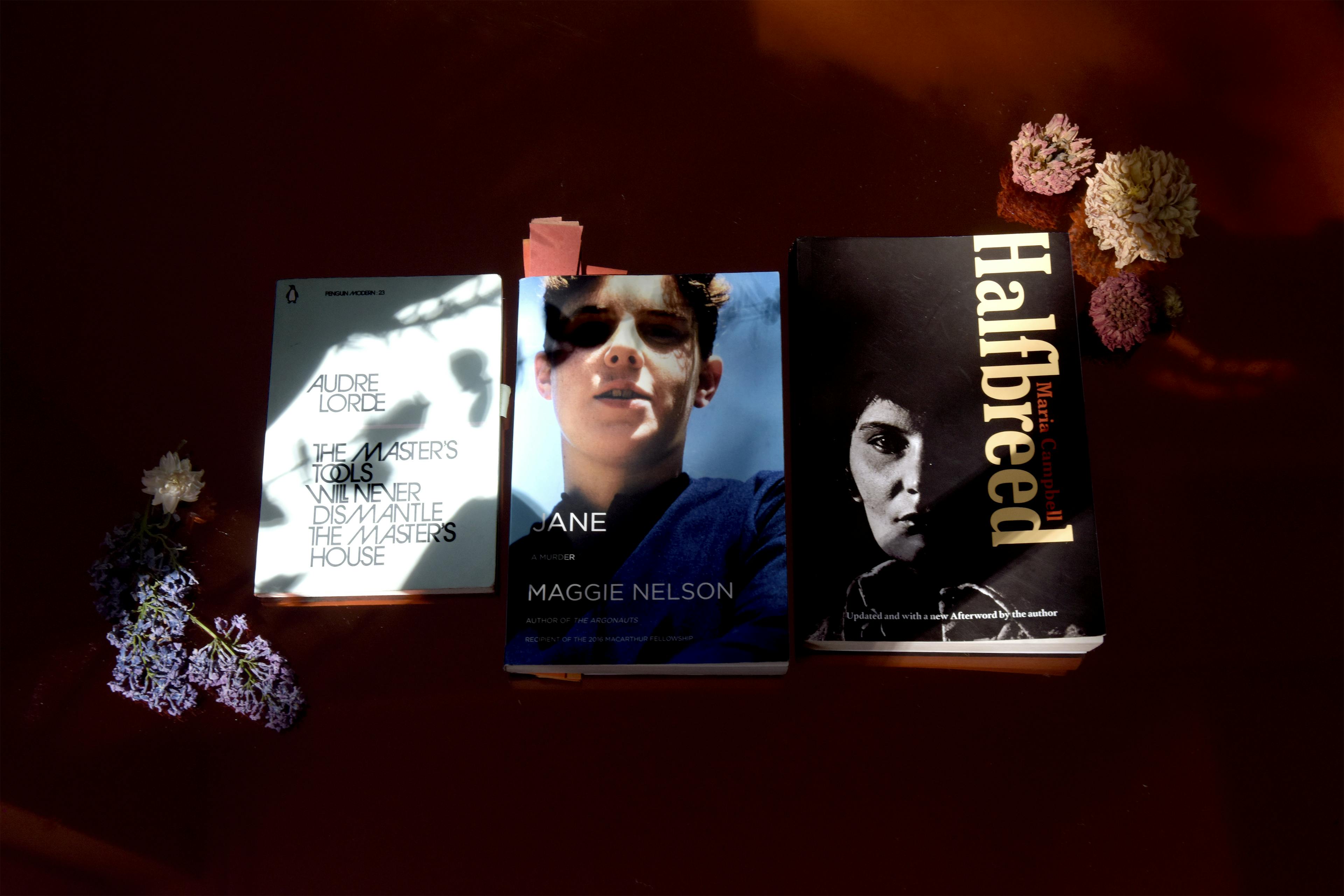 Three books are placed on a red flat surface. From the left to the right, Audre Lorde’s Poetry is Not a Luxury, Maggie Nelson’s Jane: A Murder, and Maria Campbell’s Halfbreed. 