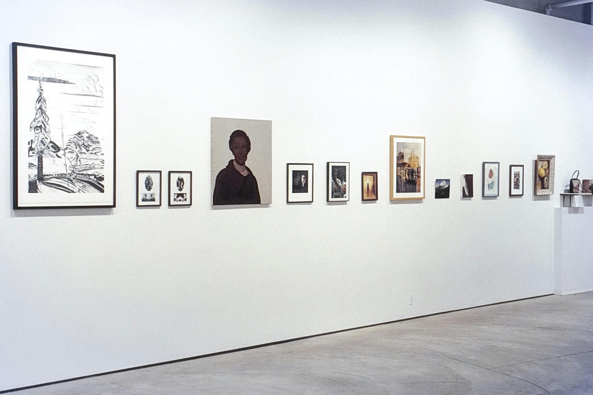 14 artworks on a gallery wall. The bottom edges of the works are mounted at the same level , but their height and width varies. The largest work, on the right, is a Salish landscape illustration.