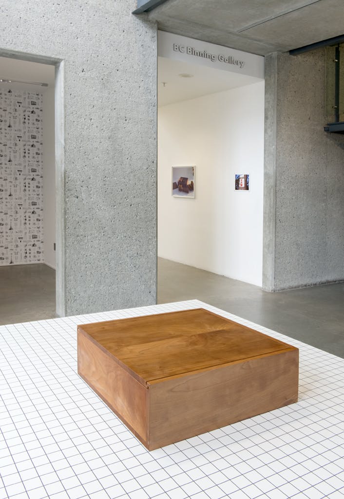 A wooden box is installed inside a gallery on a white plinth with black grids on the surface. The plinth is located in the entrance lobby. The box is fully closed. 