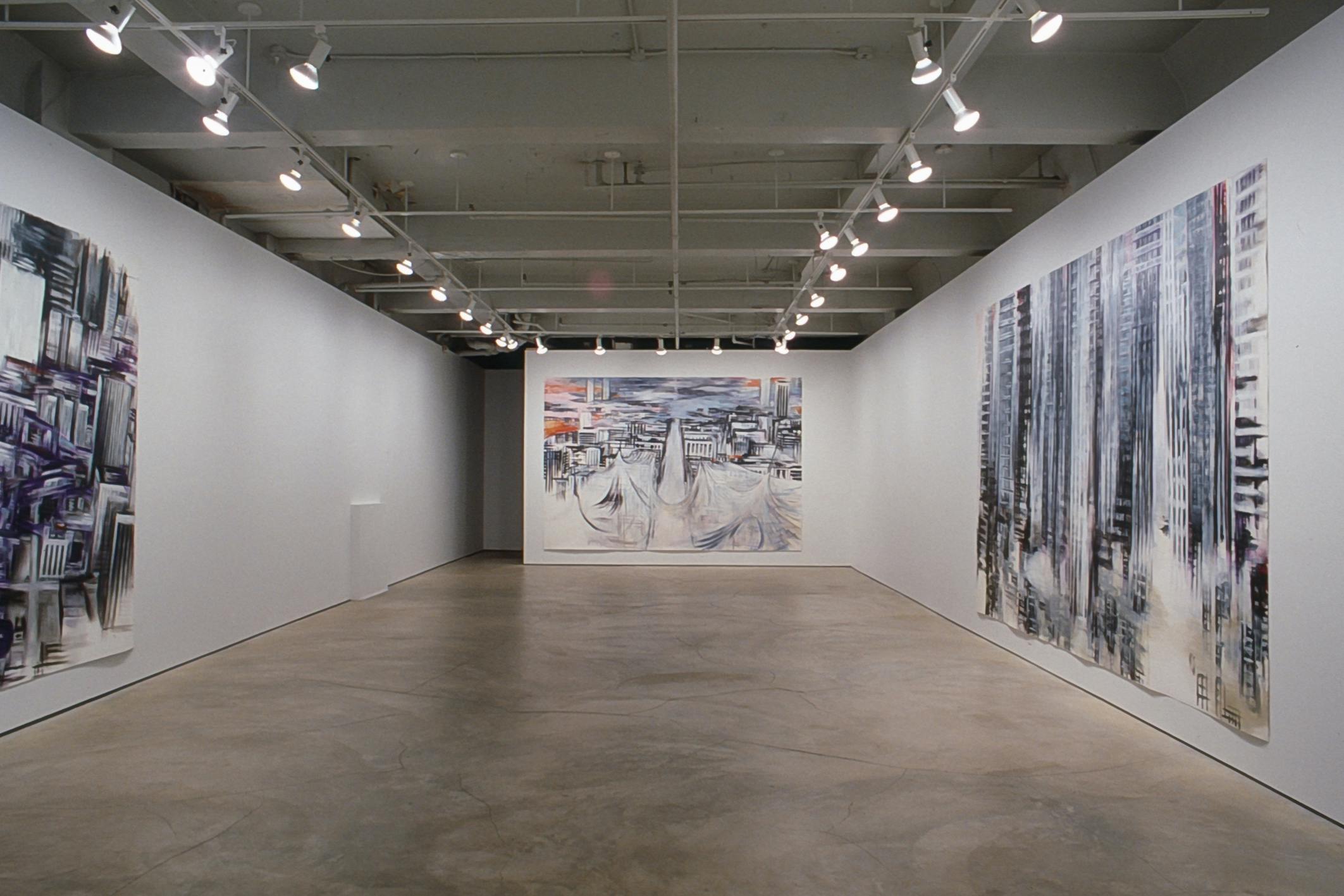 Large-scale paintings are installed on the gallery walls. They depict urban city landscapes in grey, purple, and orange colours. Many vertical lines are used to depict towers, streets and buildings.