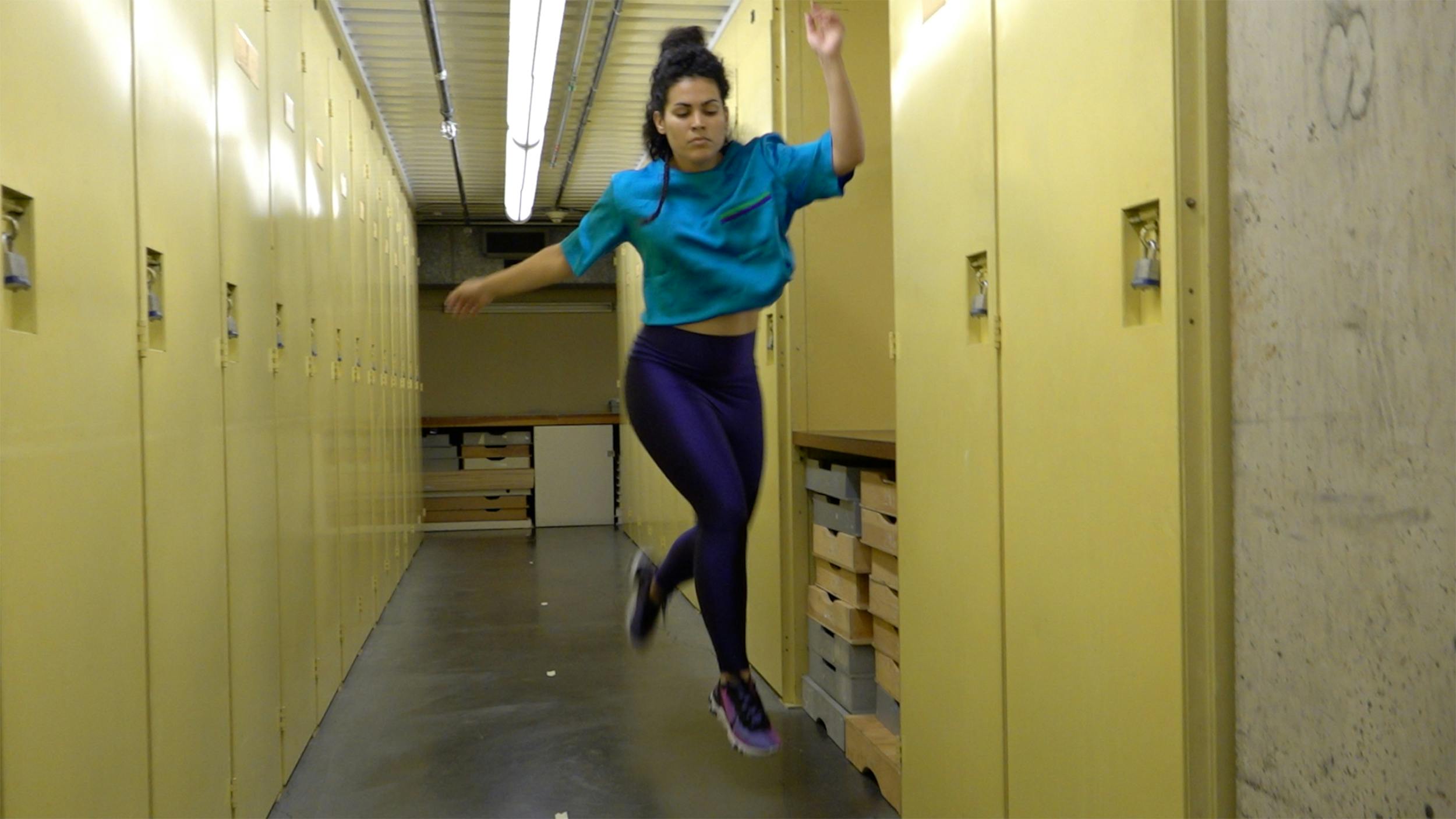 A video still of a dancer in mid-air, performing in a storage space with yellow cabinets lining the walls. She is wearing a blue shirt and purple tights.