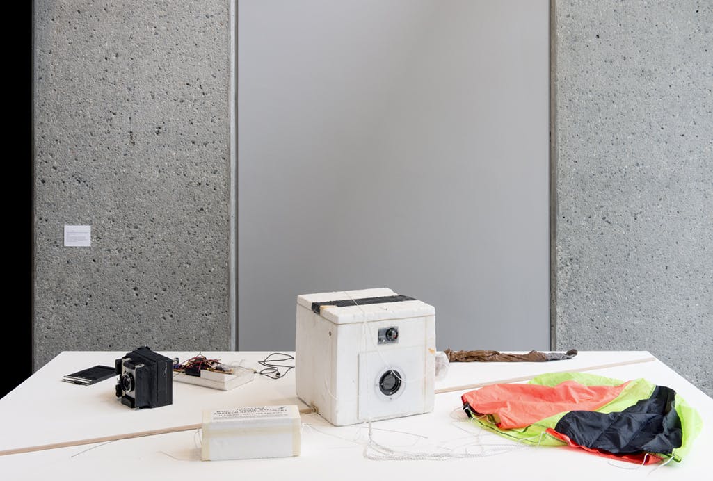 A white box with small lenses embedded on its side sits in the middle of a white surface. Other objects surround the white box: a black polaroid camera, cords, a wooden dowel, a piece of fabric.