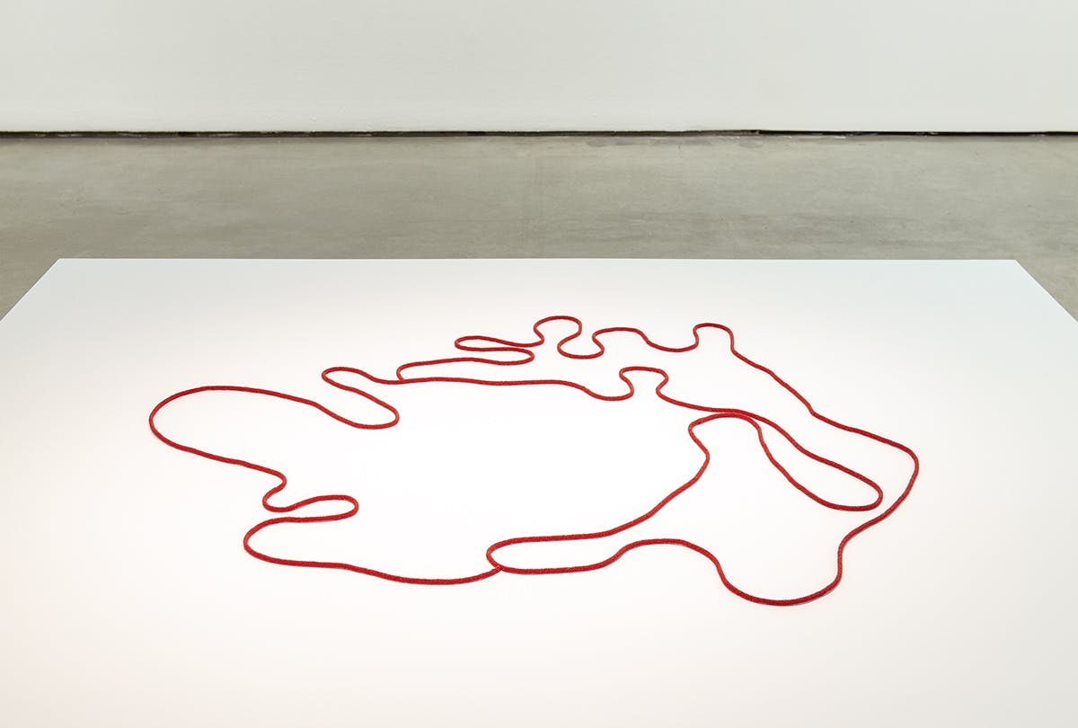 A ropelike sculpture is installed in a gallery space. The sculpture is placed on a large plinth. It is a red beaded cord forming a continuous line, maplike in shape.