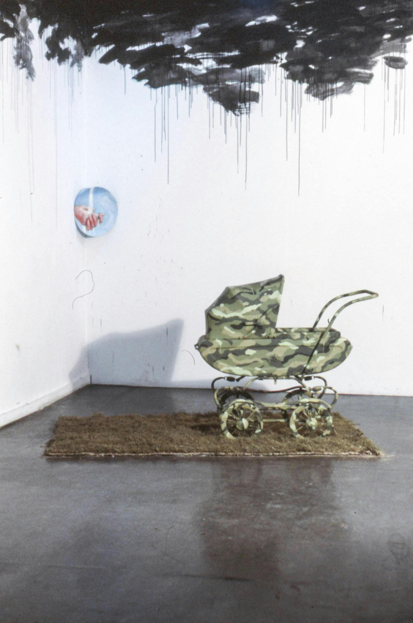 The corner of a gallery with black marks of paint at the top. Where the walls meet is a small painting of hands and a blue sky. On the floor, there is a patch of grass with a camo-print stroller on it.