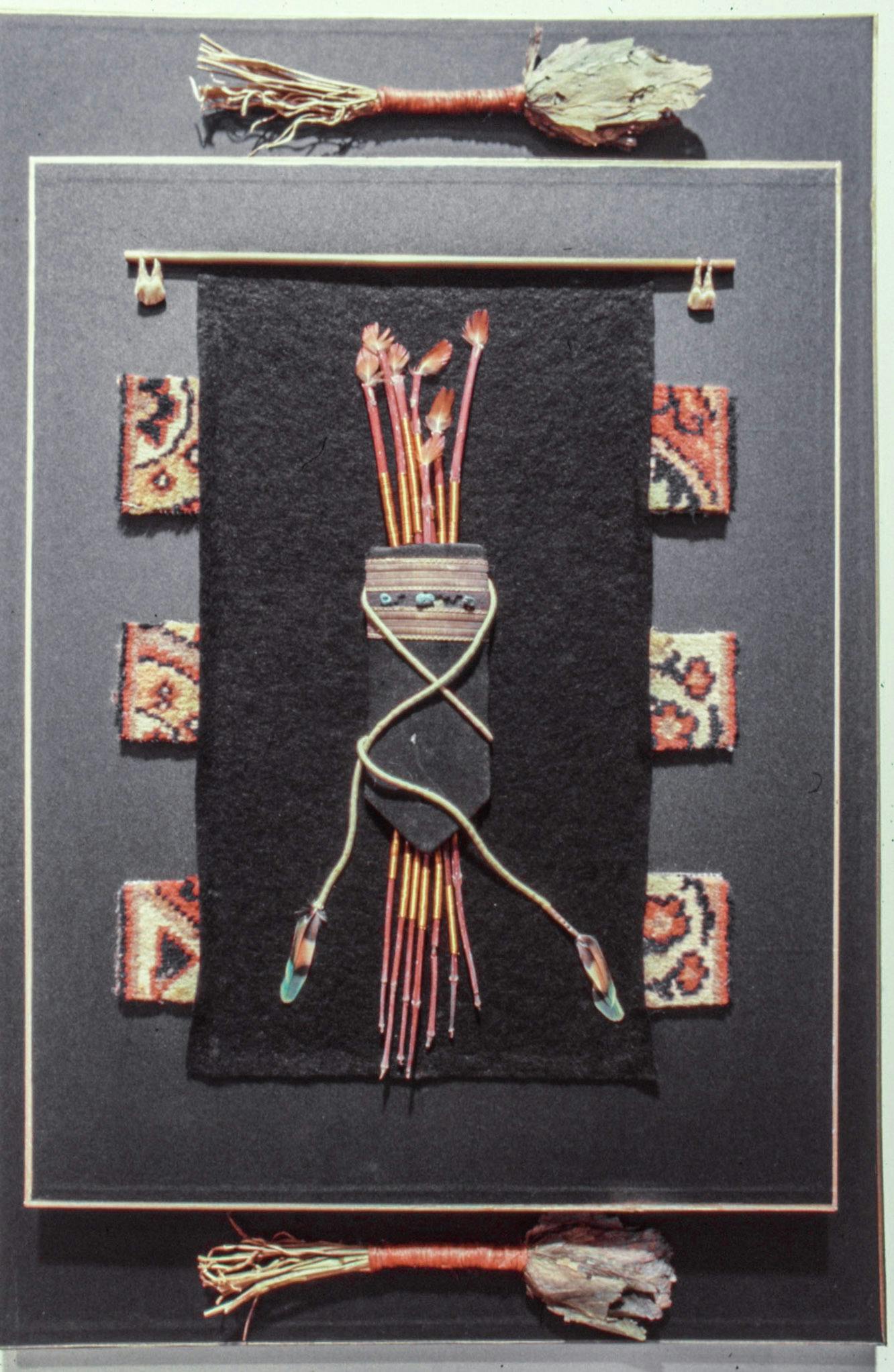 A closeup of an artwork on a wall. The work is made of several pieces mounted on black paper, including bundled dry herbs, small weavings, twined feathers and beaded leather. 