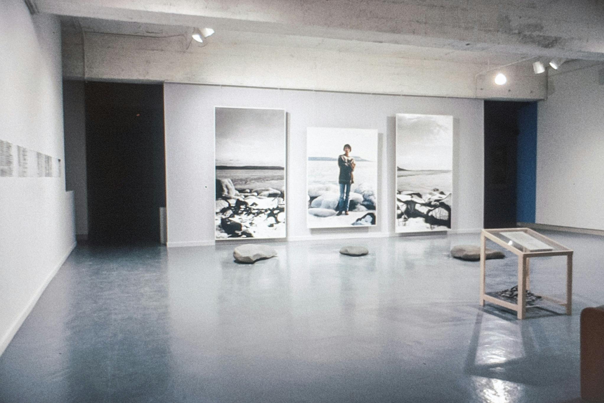 A gallery with several artworks on the walls and floor, including 3 life-scale photos. A colour photo shows a person standing on a rocky shore, dressed warmly. The other 2 show the rest of the shore.