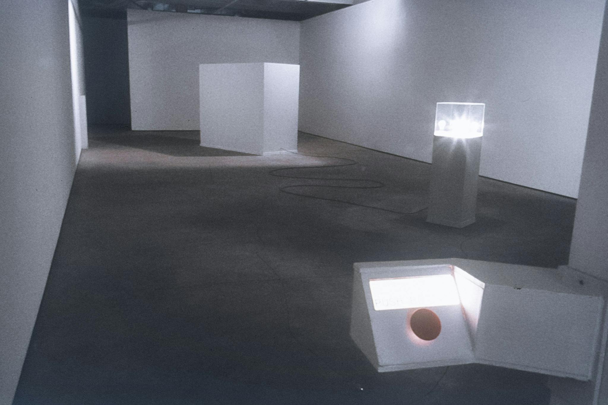 A gallery space with a button in its entryway, and two large white plinths. One plinth has a lightbulb in a glass case, and the other contains an artist, pedaling to power the lightbulb, unseen.