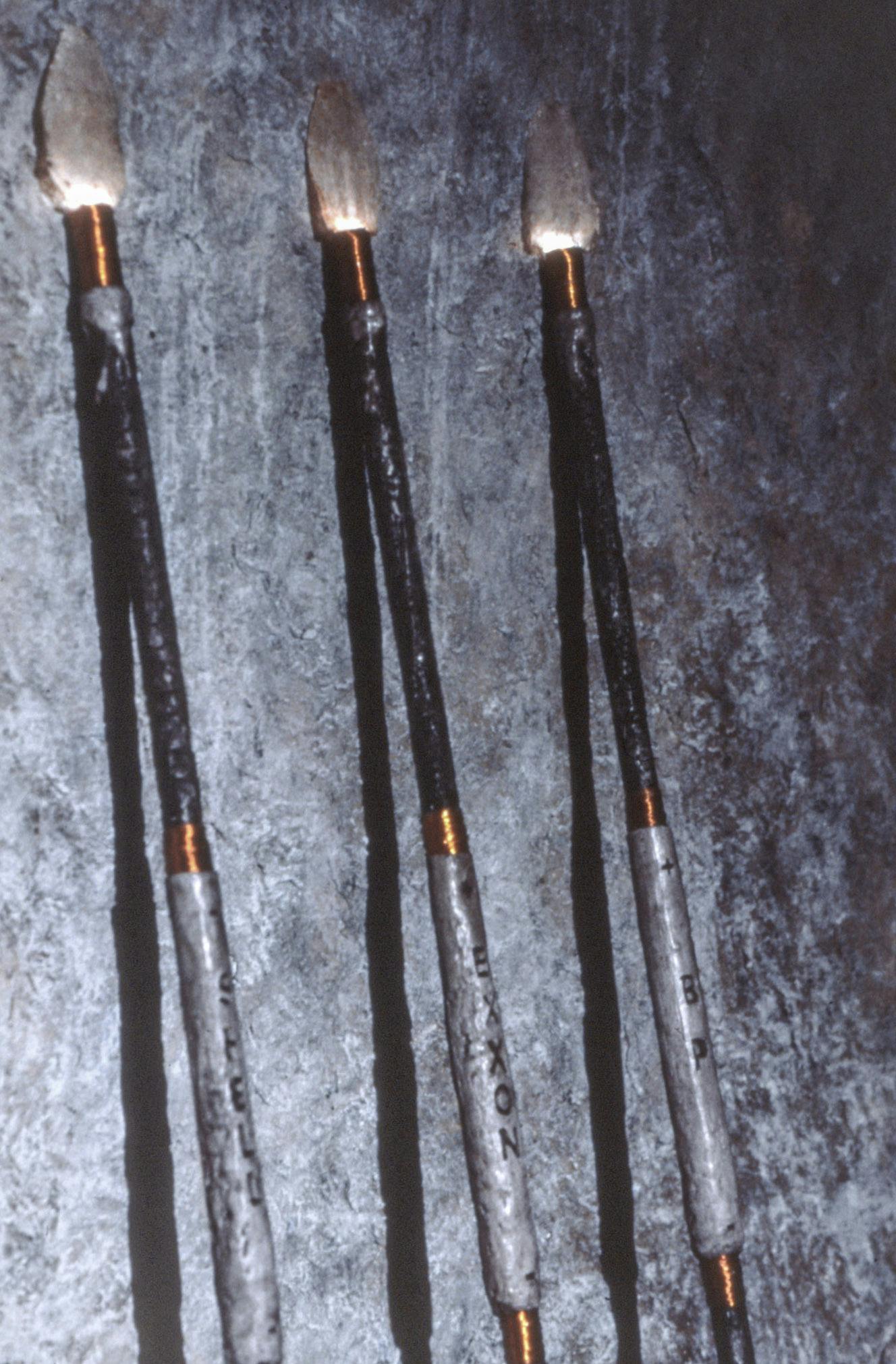 A closeup of 3 spears resting against a rock sculpture. Their points are a pearly white, attached by orange wire. On their hilts, the spears have rock texture reading "Shell," "Exxon," and "BP." 