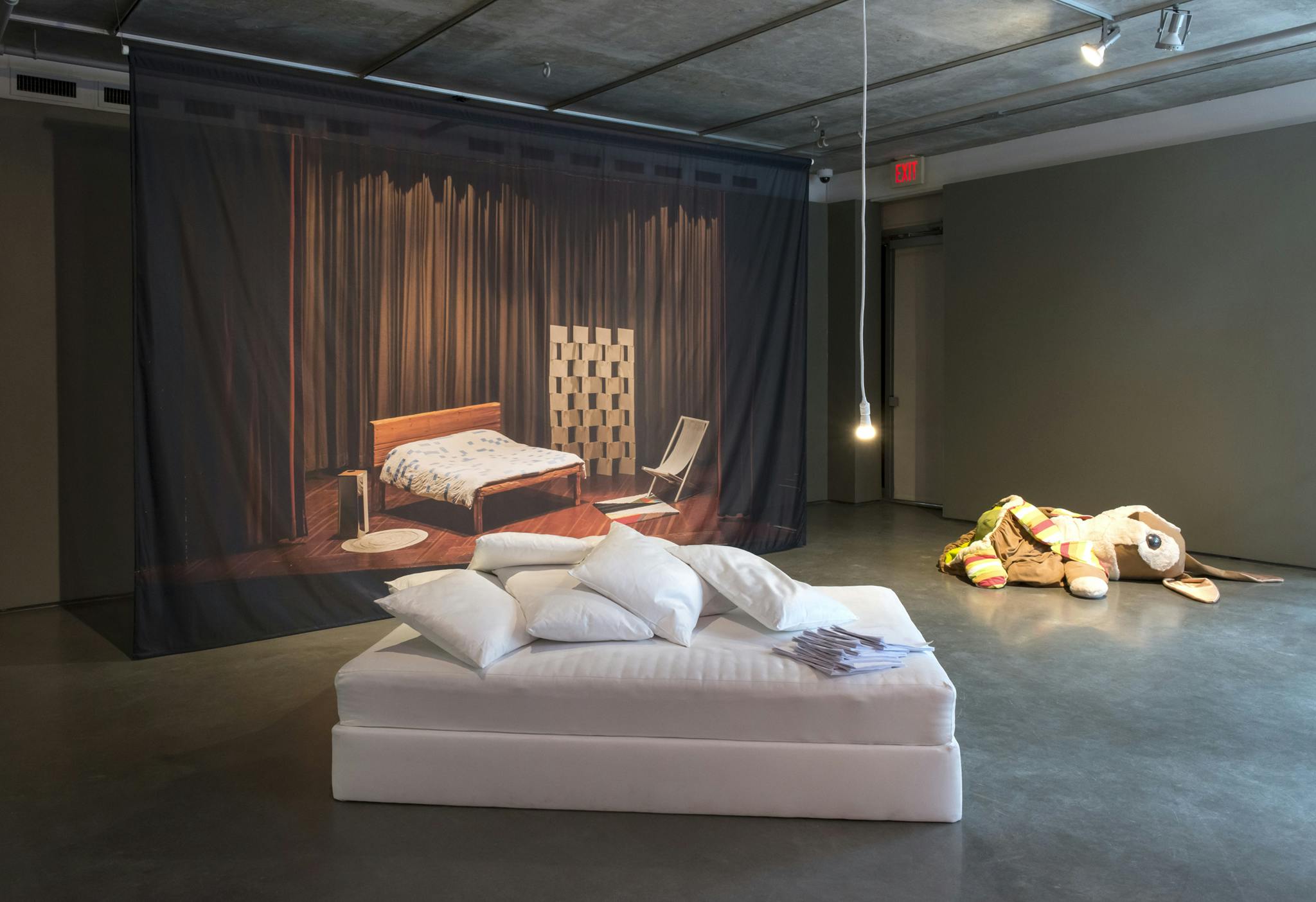 In the center of a gallery are mattresses piled with pillows. Behind a curtain with a printed photo of a bedroom stage set hangs from the ceiling and a large stuffed animal bunny lies on the floor. 