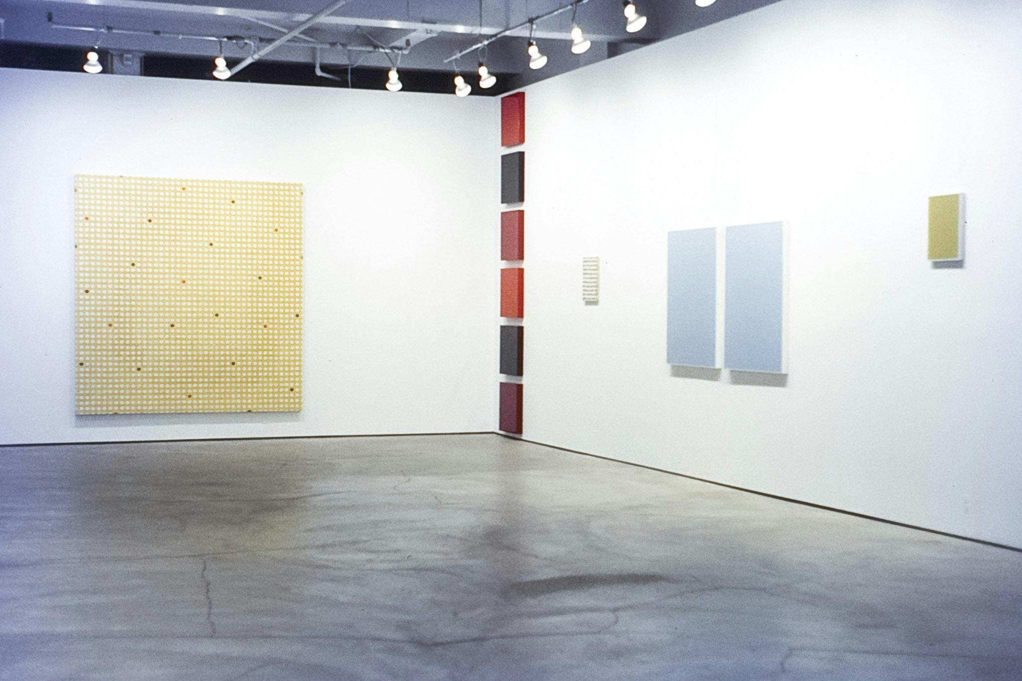 The corner of a gallery space with various artworks mounted on both walls. The works are paintings on square/rectangle canvasses. Many of them are one single colour but two have a dotted pattern. 