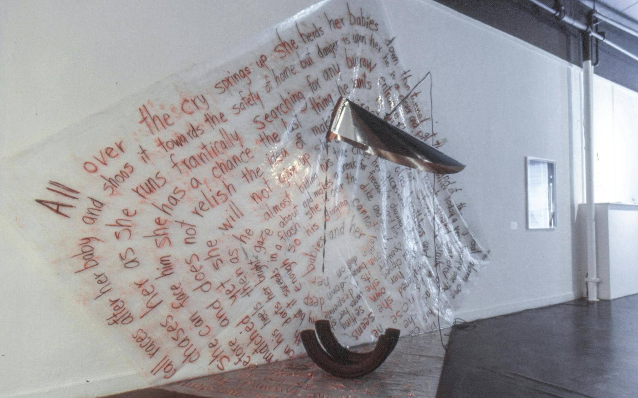 A closeup of an artwork. The work is a large plastic sheet with text that is on both the wall and the floor. There are two rusty curved pipes on the floor, and a folded silver panel on the wall.