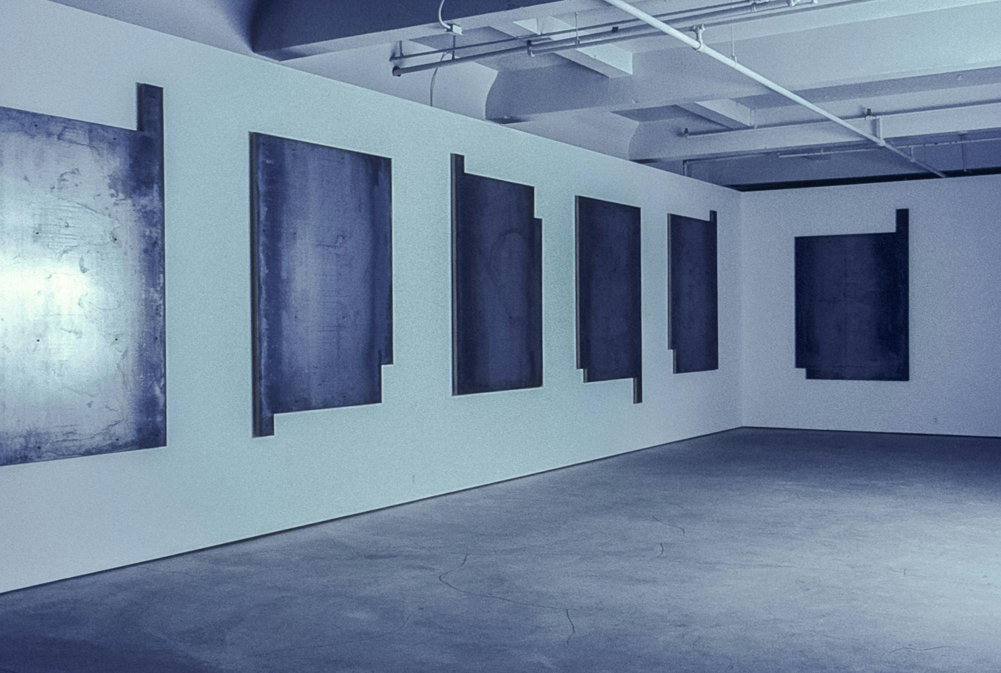 In the corner of a gallery, several rectangular metal panels hang on the wall. They have small rectangular pieces missing and protruding from their corners. Their surfaces are scuffed and marked.