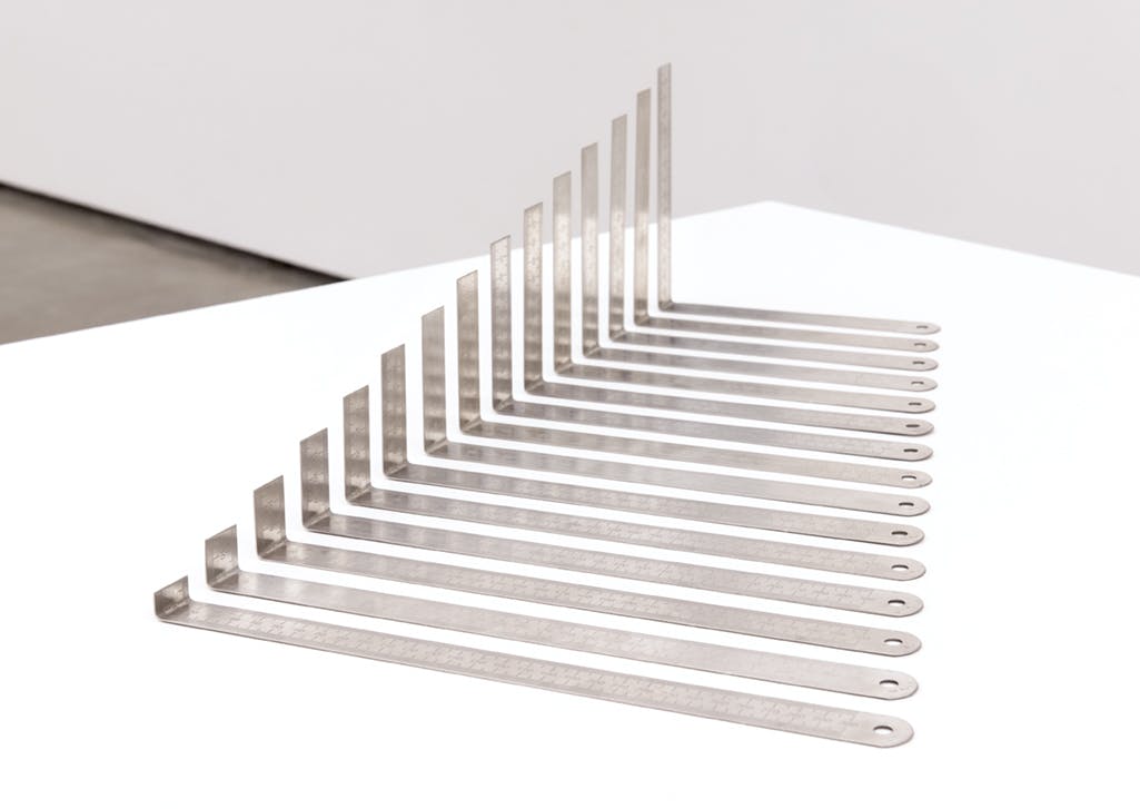 Fifteen metal rulers align diagonally on a white table in a gallery. Each is bent upwards at a 90 degree angle at different points. In combination, the aligned bent sections create a raised slope.