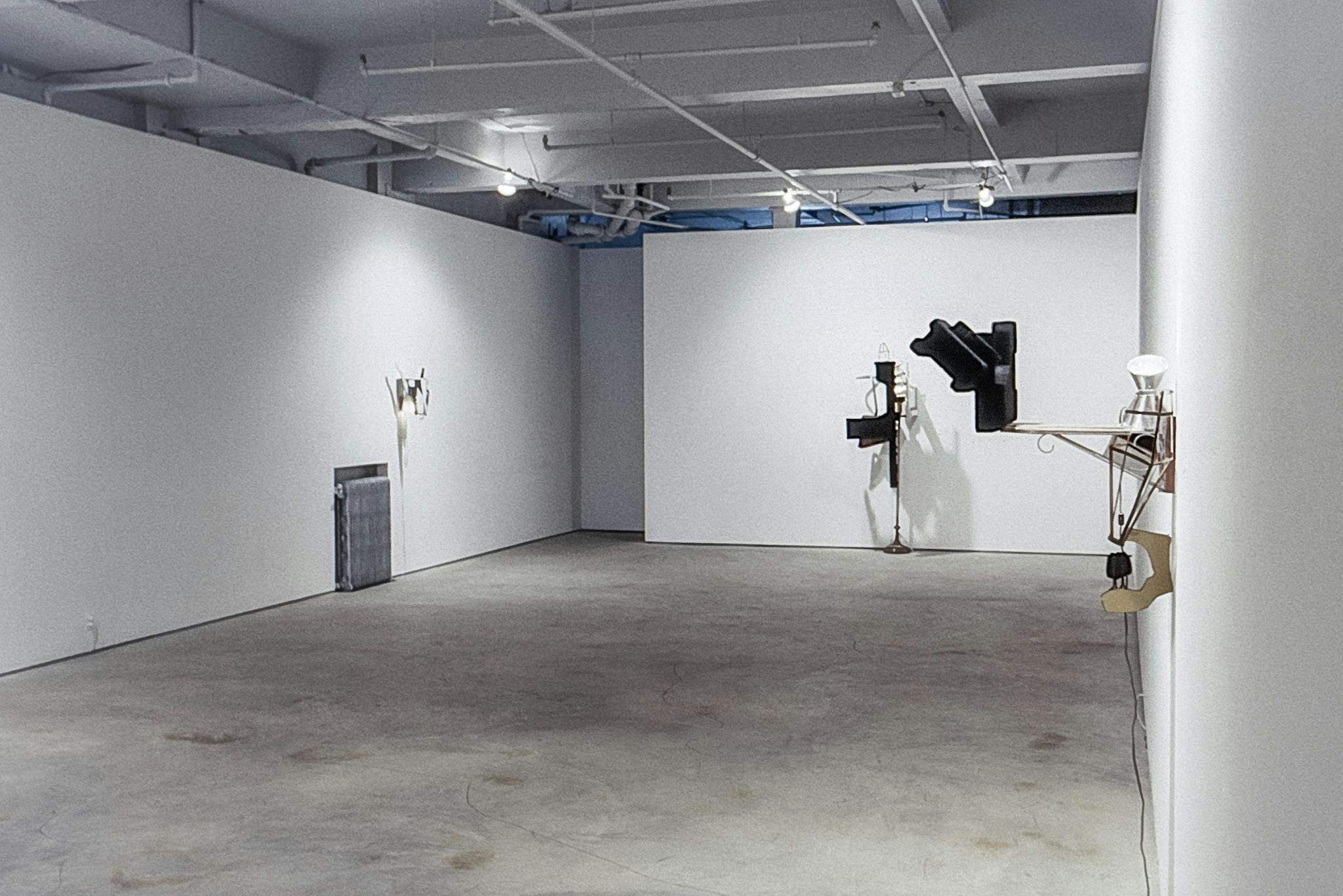 A gallery space with 3 small sculptures on the walls. They all have different metal elements, including a plant hood, a lamp base, and metal sheets that are cut and bent. 