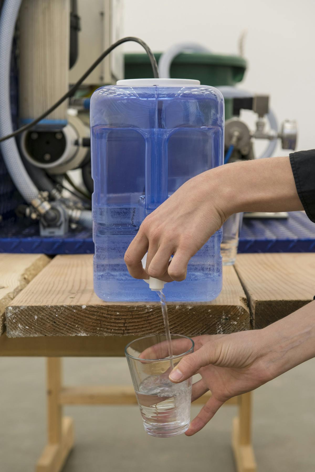 A blue water jug sits on a wood table, from which a pair of hands pour water into a glass. A machine sits behind the tank. This machine and the tank are connected by a black tube. 