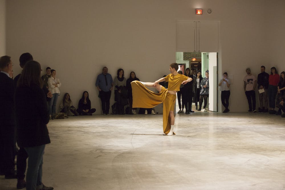 A gallery space with people standing around the perimeter of the room watching one person perform at the center. The performer is holding their leg up at a ninety degree angle in yellow loose clothes. 