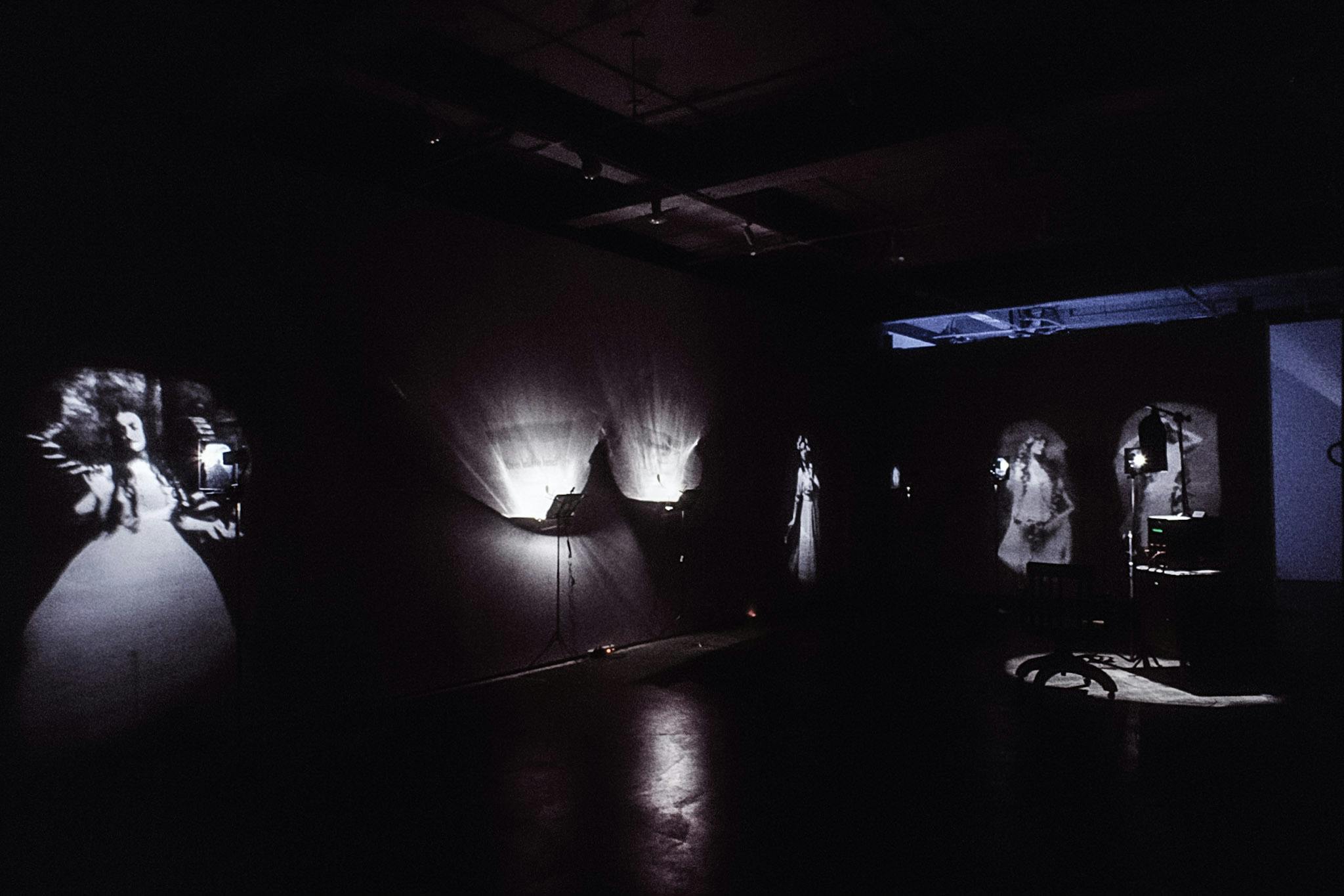 An installation image of a dimly-lit gallery. A pair of stage lights are installed closely to each other near the middle of a wall. Several black and white images of women in dress are projected on the walls.