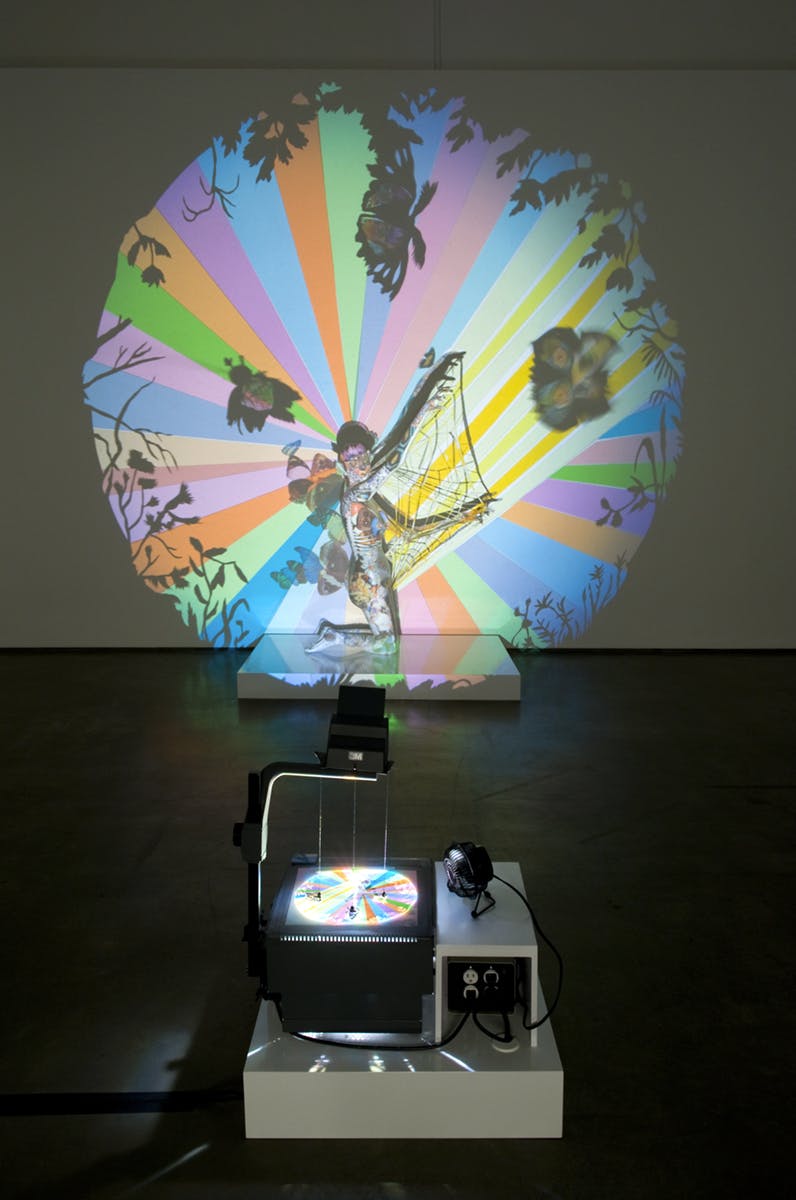 An overhead projector points towards a wall, casting a colourful projection onto a human-like sculpture posing on a platform. The projection is a circular array of stripes, butterflies, and tree shadows.