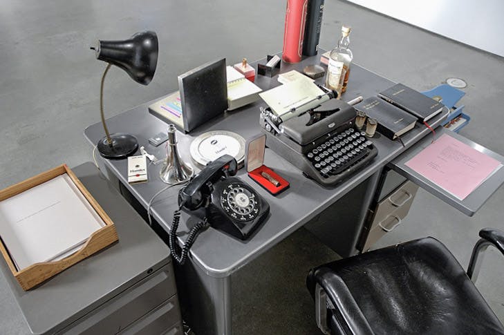An installation image of a work by Daniel Olson. A steel-made grey working desk and a black leather-covered office chair are placed on the floor. The desk is covered with various objects, including a typewriter, a desk lamp, and a cigarette case.