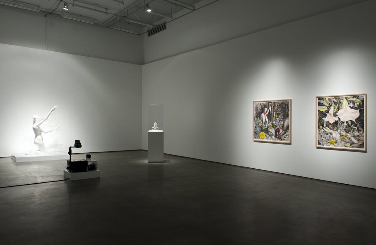Two large-scale artworks are mounted on a gallery wall. On the floor are two white sculptures, one a large, human-like form of white plaster and another, a smaller sculpture under glass atop a plinth.