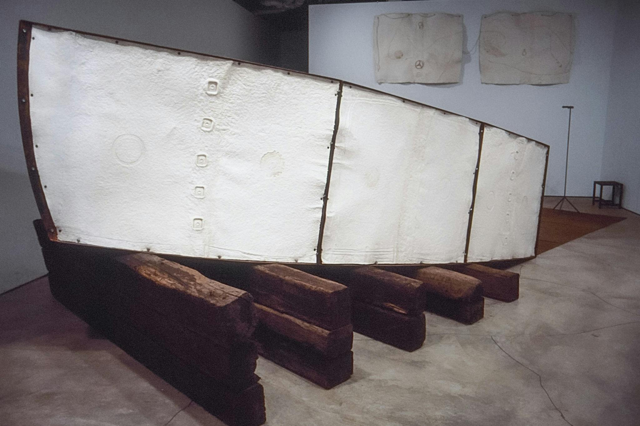 This is a side view of a boat head sculpture. White panels that seem to be made of parchments or pulp sheets cover the body of the boat. The boat is placed on the wooden bars placed on the floor. 