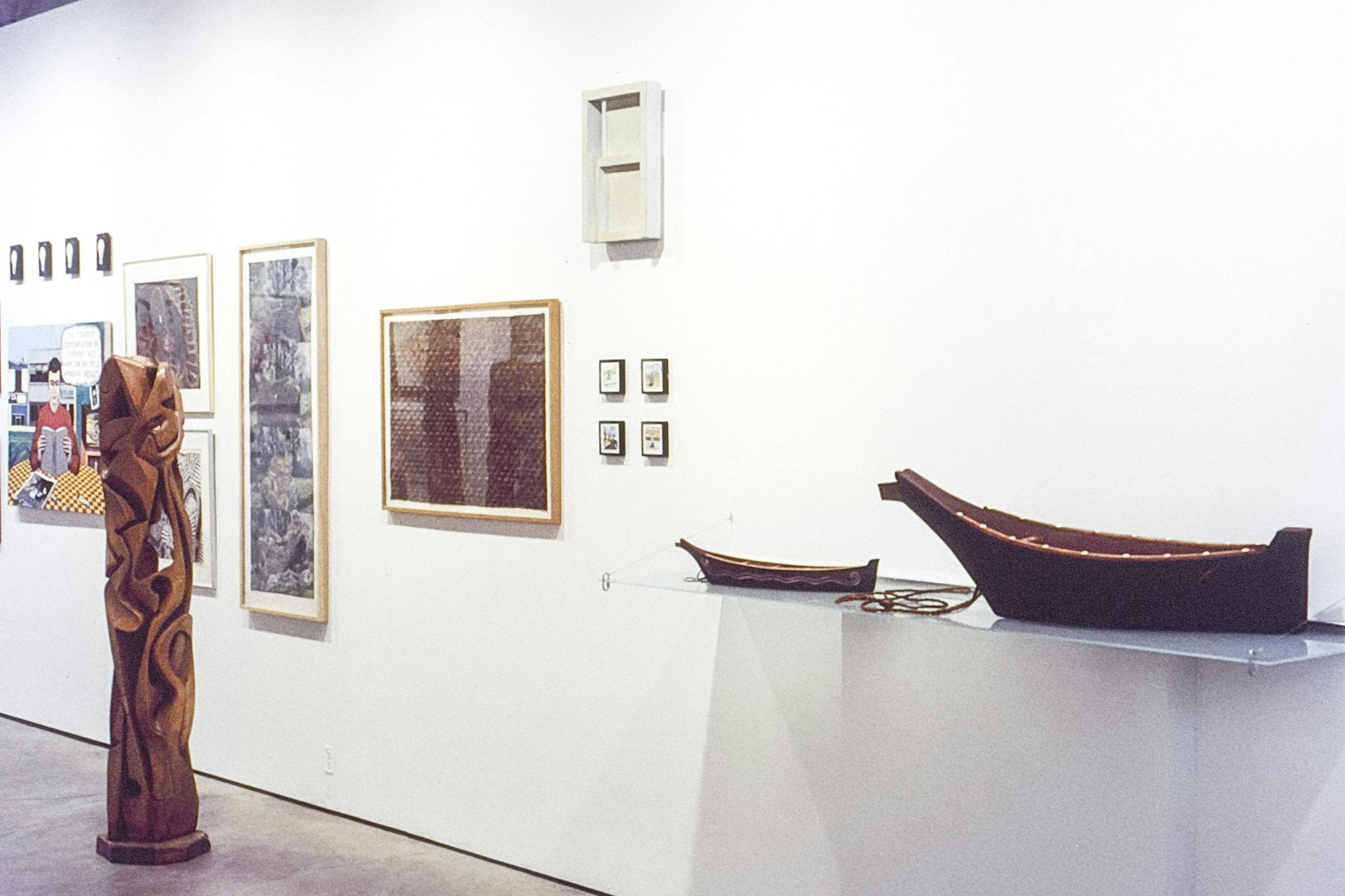 A photo of artworks installed in the gallery. A set of small-scaled two black canoes are placed closer to the camera; one is bigger than the other, and the smaller one has a snake drawn on its side. 