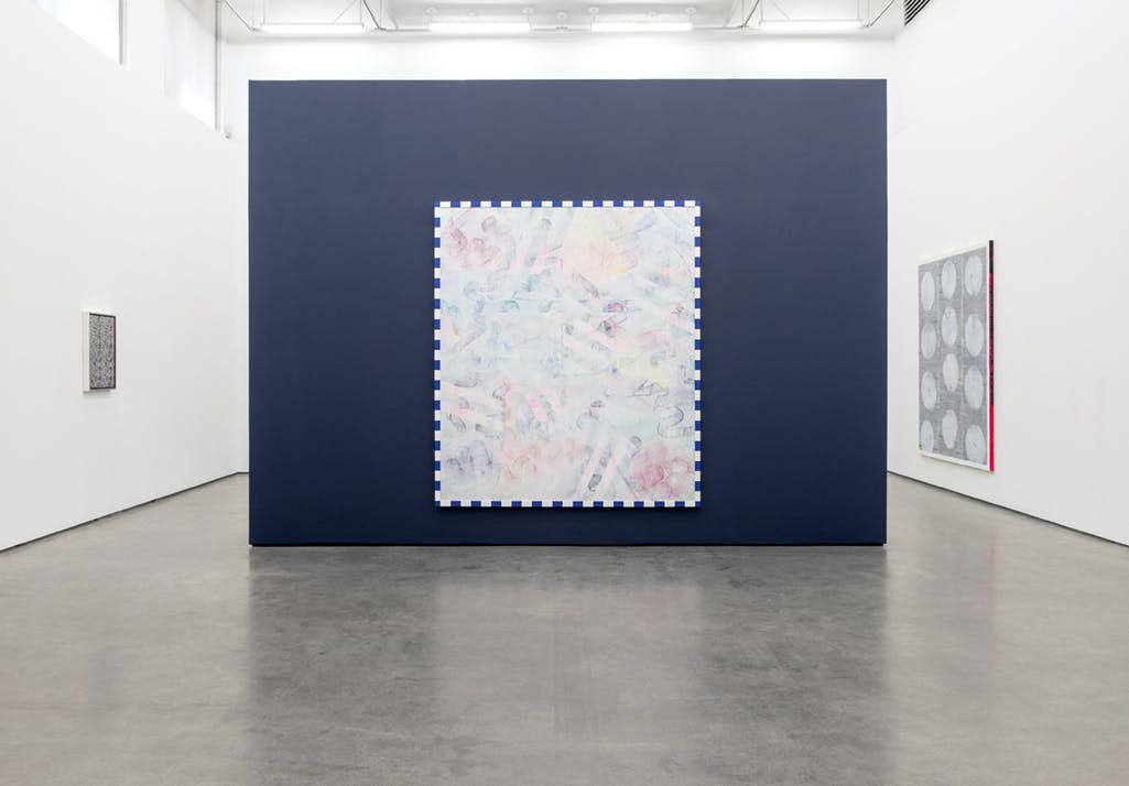 A large-scale painting is installed on a blue, free-standing wall. In light tones, abstract shapes and colours repeat across the canvas. Alternating blue and white rectangles frame the edge.