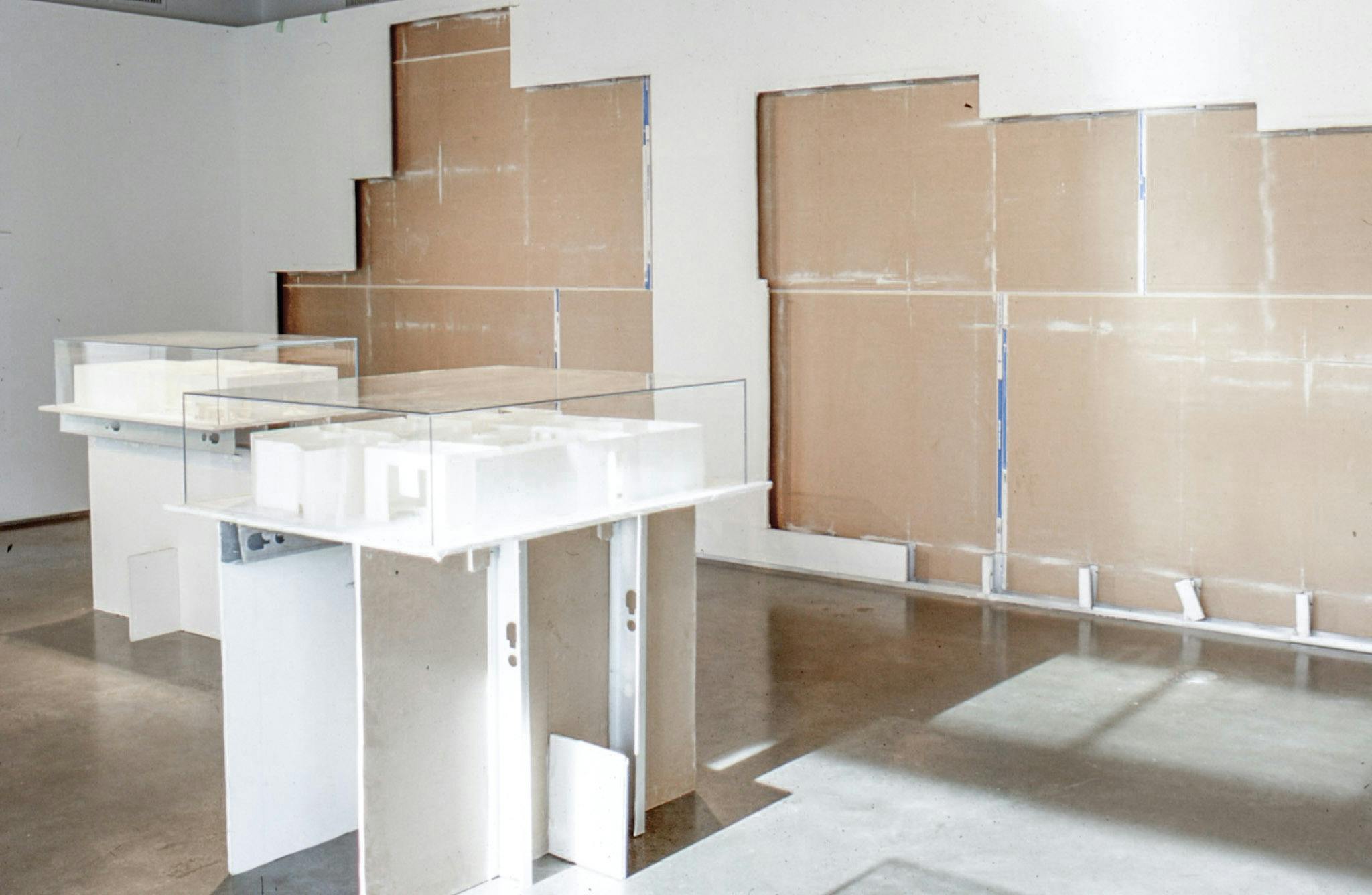 A set of white sculptures in display boxes are placed in a gallery space. White tiles on the gallery wall were removed, making the brown internal part of the wall visible. 
