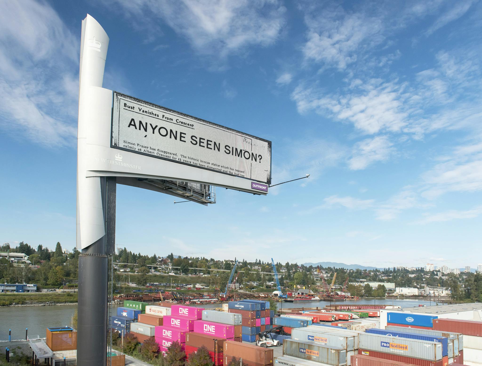 A billboard stands high off the ground with a bridge on one side and shipping containers on the other. The billboard reads “Anyone Seen Simon?” in black text. 