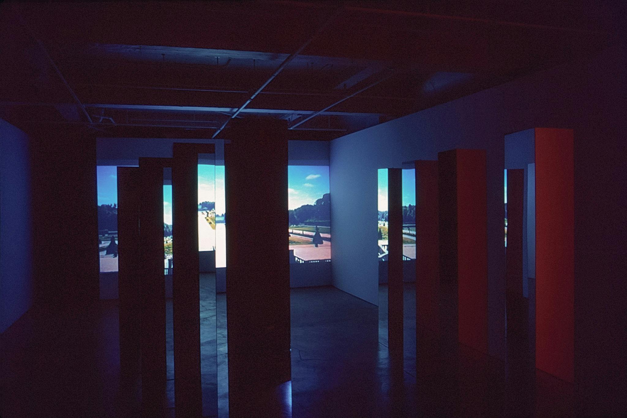 Eight large mirrored columns stand on the gallery floor. A photographed image of a garden is projected on the front wall. Reflections on the mirrors show a red light illuminating the back wall.