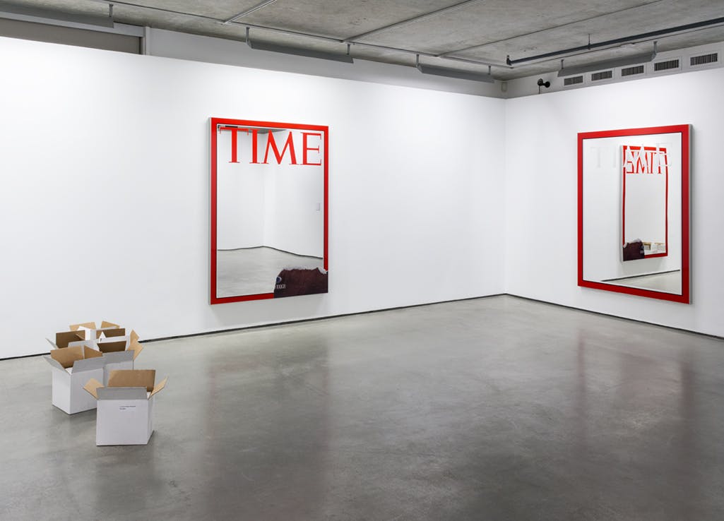 A pair of large-scale mirrors are installed on white gallery walls. They bear the logo and red border of TIME Magazine. Five white cardboard boxes are grouped on the floor. 