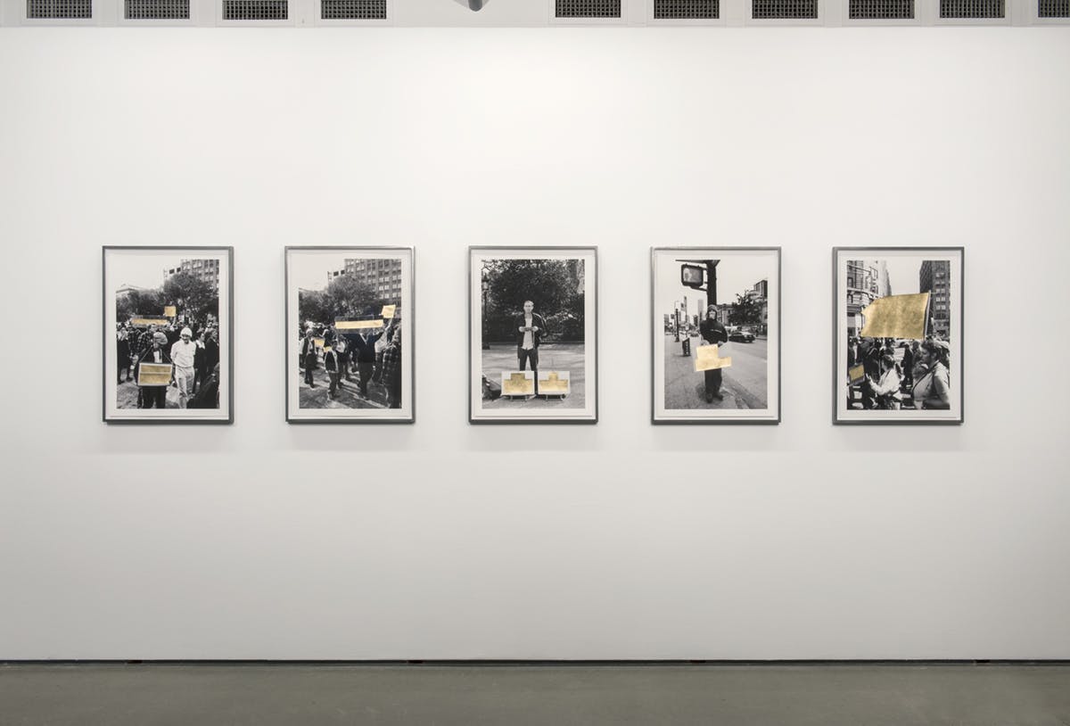 A series of five framed photographs are mounted on a gallery wall. Each of them shows different scenes from a street protest. Flags and signs are covered by an intervention of gold leaf over the image.