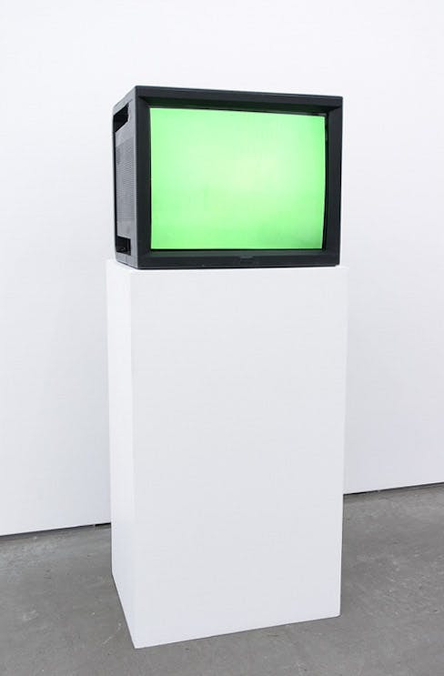 An installation view of a work titled Watercolour by Ceal Floyer. A CRT TV installed on a white pedestal displays a surface of a white screen illuminated in green.